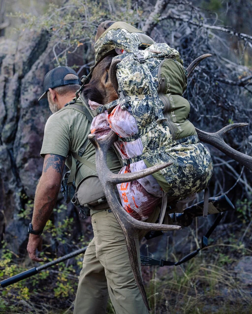 Ein Mann in Outdoor-Ausrüstung wandert mit einem EBERLESTOCK Vapor 5000 V2 Rucksack, der mit Elchgeweih und Fleisch beladen ist, durch den rauen Wald.