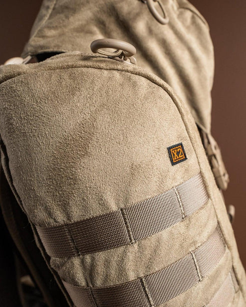 Nahaufnahme des EBERLESTOCK X2 - Mountain, eines braunen Wildlederrucksacks mit horizontalen Gurtbändern und einem kleinen rechteckigen Logoaufnäher auf der Vorderseite, der ein Volumen von 40 Litern bietet, vor einem braunen Hintergrund.