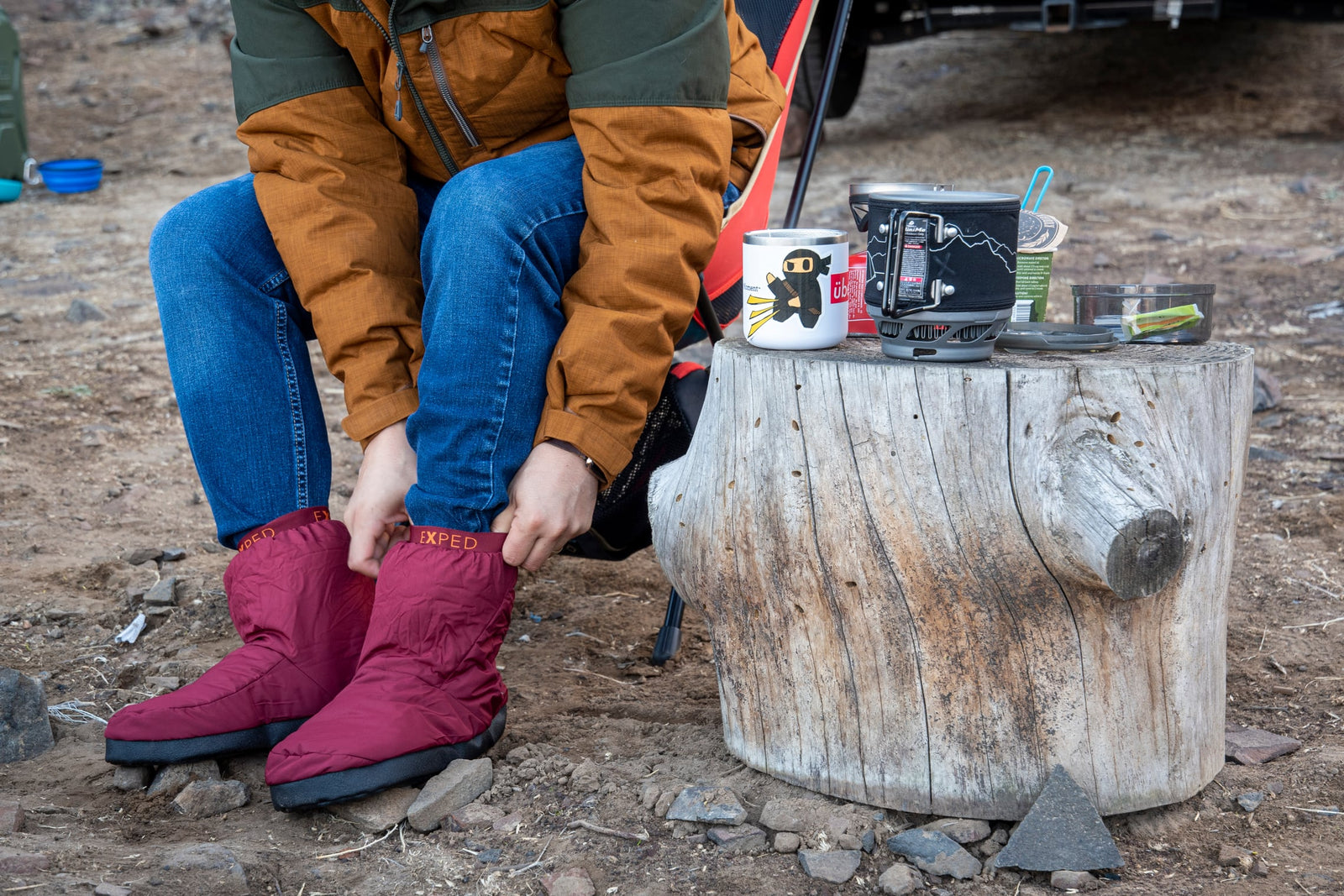 Eine Person in einer braunen Jacke und blauen Jeans sitzt im Freien und zieht die Camp Booty - Navy mit rutschfester Sohle von EXPED an. In der Nähe stehen auf einem Baumstumpf eine Tasse, ein Campingkocher und andere kleine Campingutensilien.