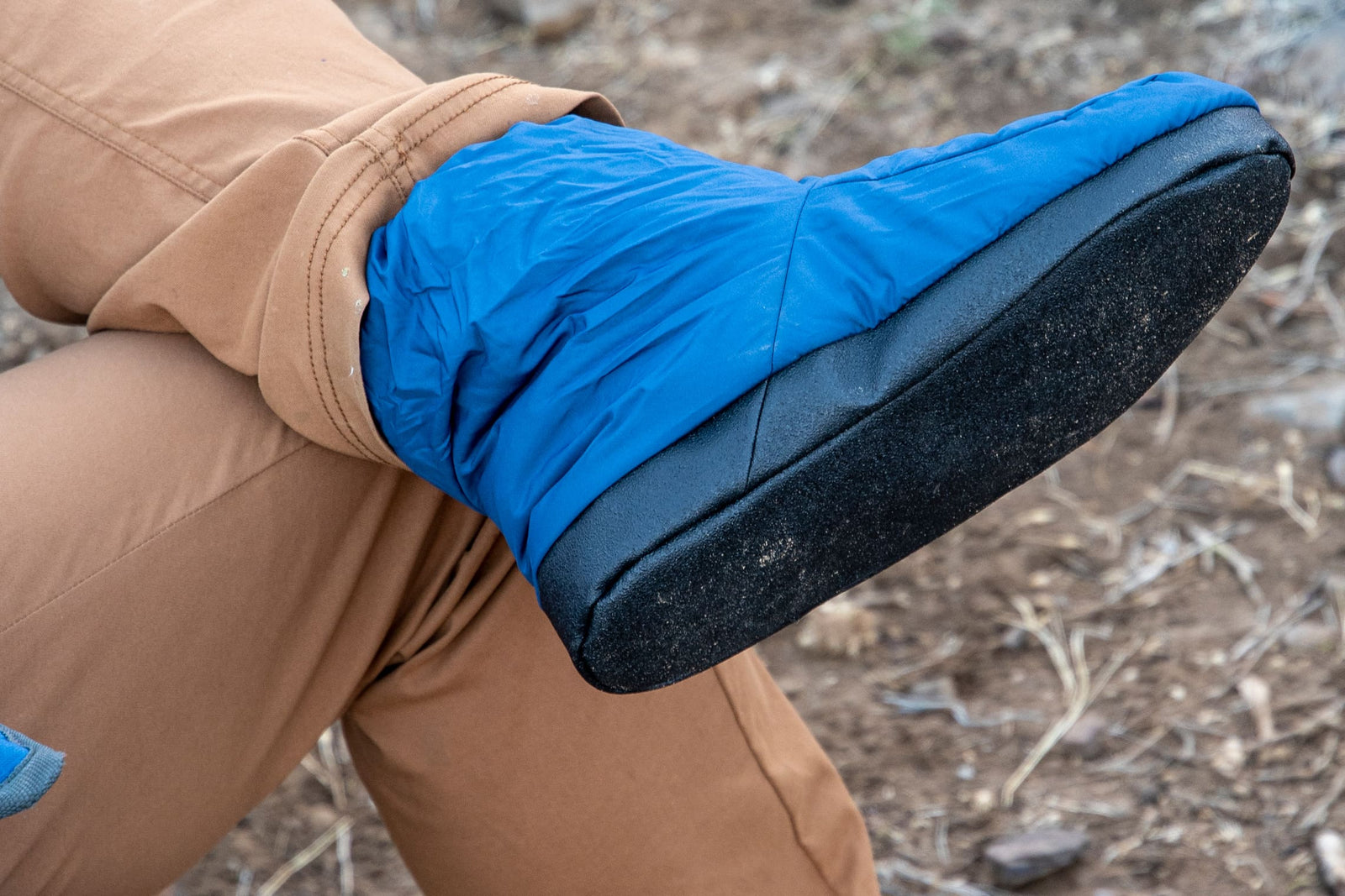 Eine Person in hellbrauner Hose und EXPED Camp Booty - Navy mit rutschfester Sohle sitzt im Freien auf trockenem, felsigem Boden. Der leichte Schuh von EXPED wird in Nahaufnahme gezeigt, wie das Bein über das Knie geht.