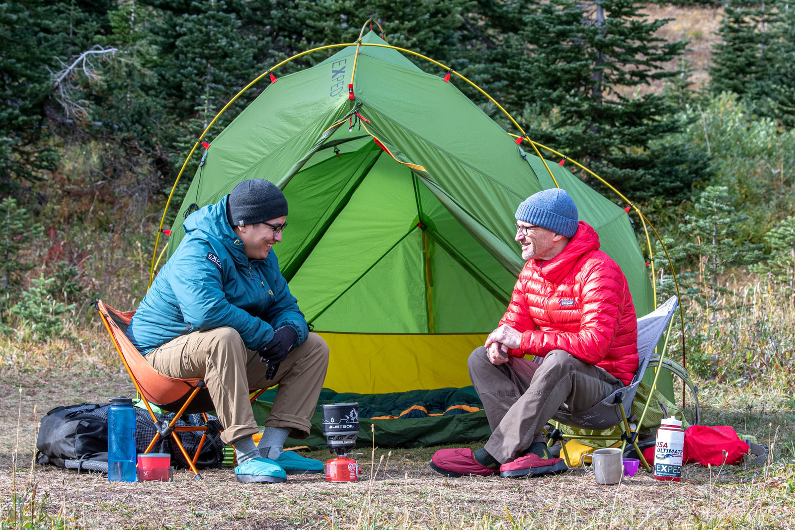 Zwei Personen in warmen Jacken und Mützen entspannen sich vor einem grünen Zelt, umgeben von Campingausrüstung. Beide tragen EXPED Camp Slipper - Navy mit rutschfester Sohle, während sie sich unterhalten, während Tassen und ein Wasserkocher im Gras stehen.