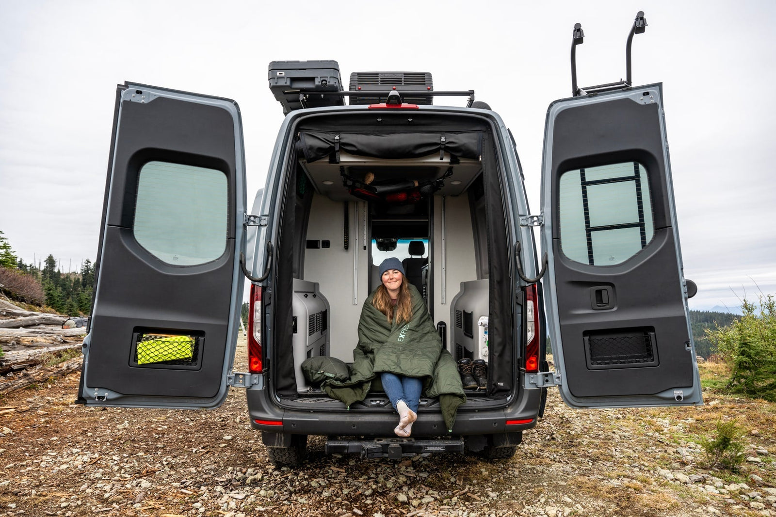 Eine Frau sitzt eingewickelt in eine EXPED Decke LuxeWool Blanket DUO auf dem Rücksitz eines offenen Wohnmobils, das im Freien auf felsigem Gelände mit Bäumen und Bergen im Rücken geparkt ist. Die Hecktüren des Vans sind weit geöffnet.