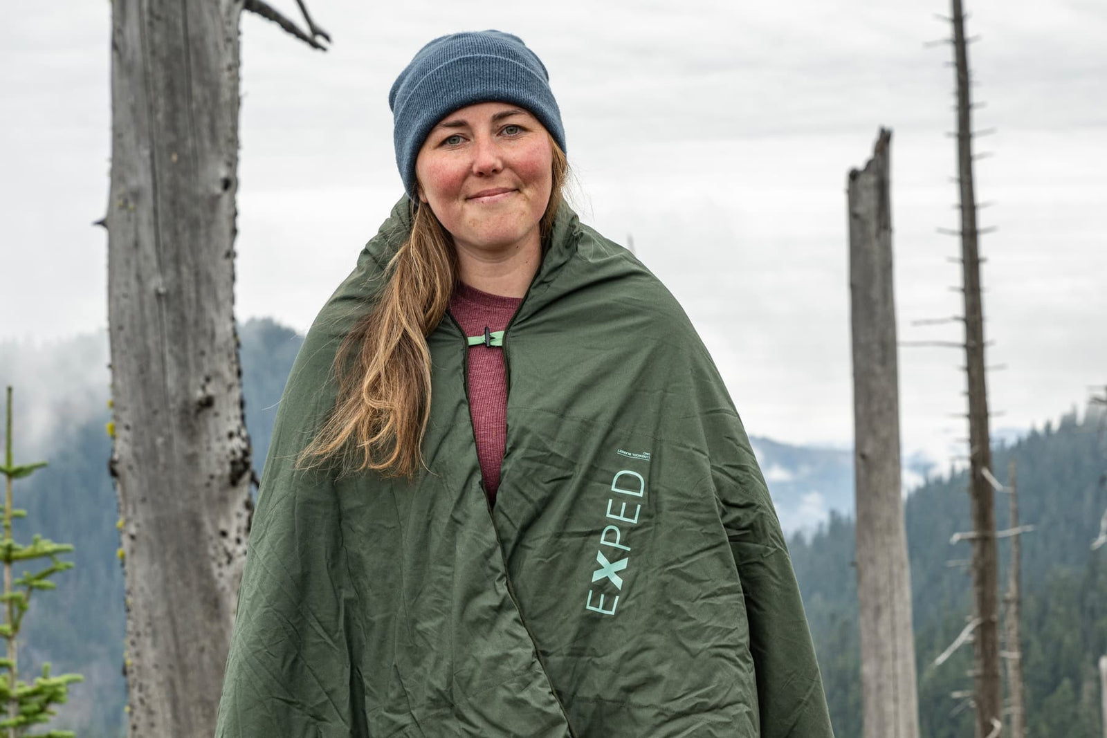 Eine Person, die eine blaue Mütze trägt und in eine olivgrüne EXPED Decke LuxeWool Blanket UNO eingewickelt ist, steht im Freien in einer bergigen, bewaldeten Gegend bei bewölktem Himmel.