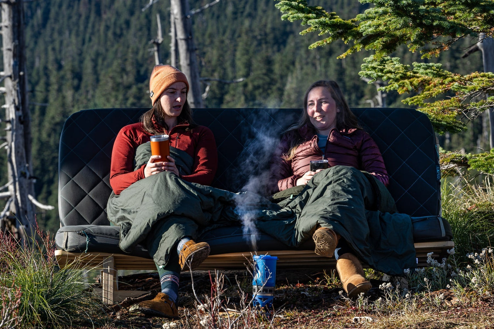 Zwei Frauen entspannen sich im Freien in Schlafsäcken, Tassen in der Hand, lächelnd. Eine EXPED Decke LuxeWool Blanket UNO bedeckt ihre Schöße, während Dampf aus einem blauen Campingkessel aufsteigt. Pinienbäume und bewaldete Berge bilden eine gemütliche Kulisse.