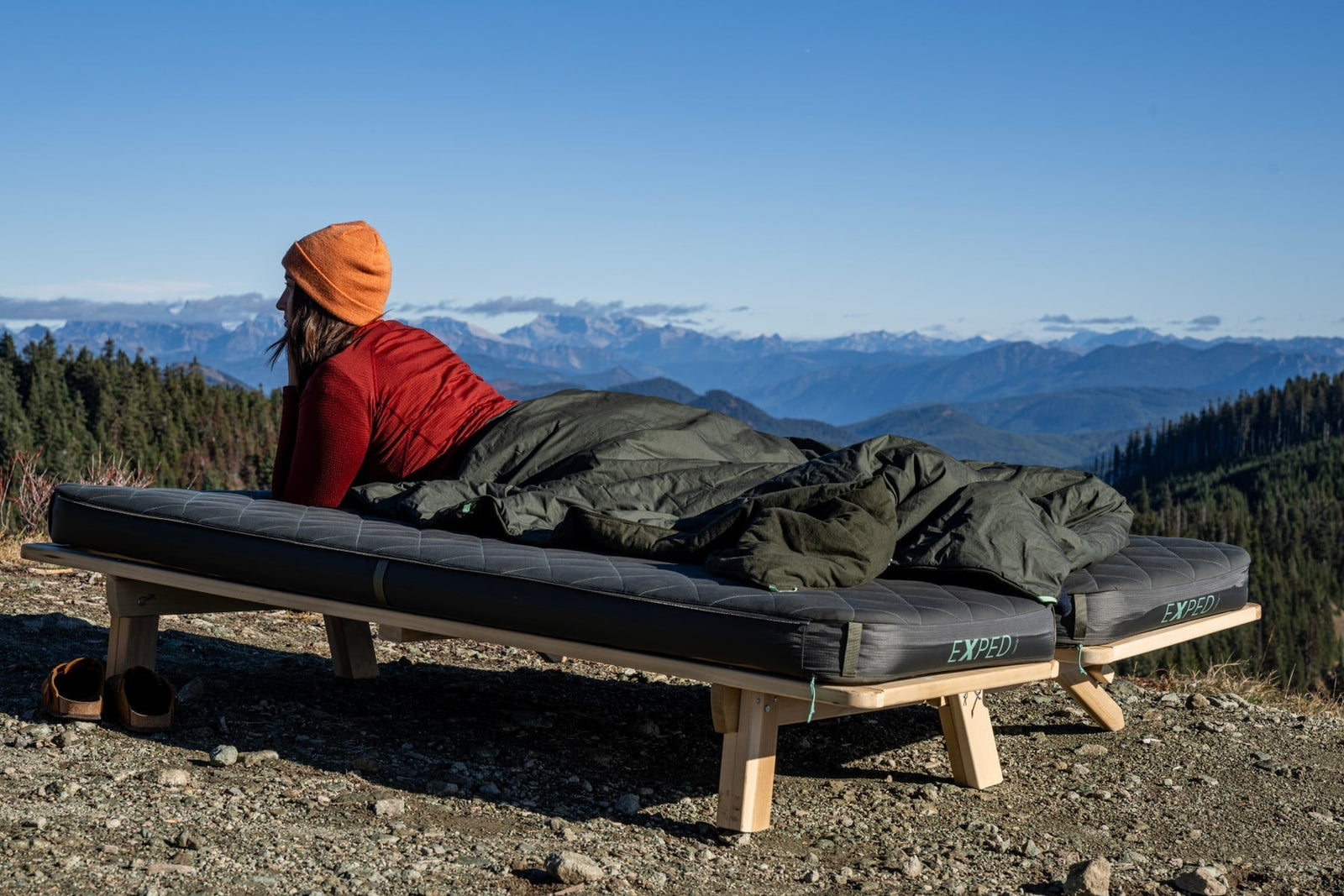 Eine Person mit rotem Pullover und orangefarbener Mütze entspannt sich auf einem Feldbett mit Schlafsack und einer kuscheligen EXPED Decke LuxeWool Blanket UNO und blickt auf die malerischen Berge unter einem klaren blauen Himmel.