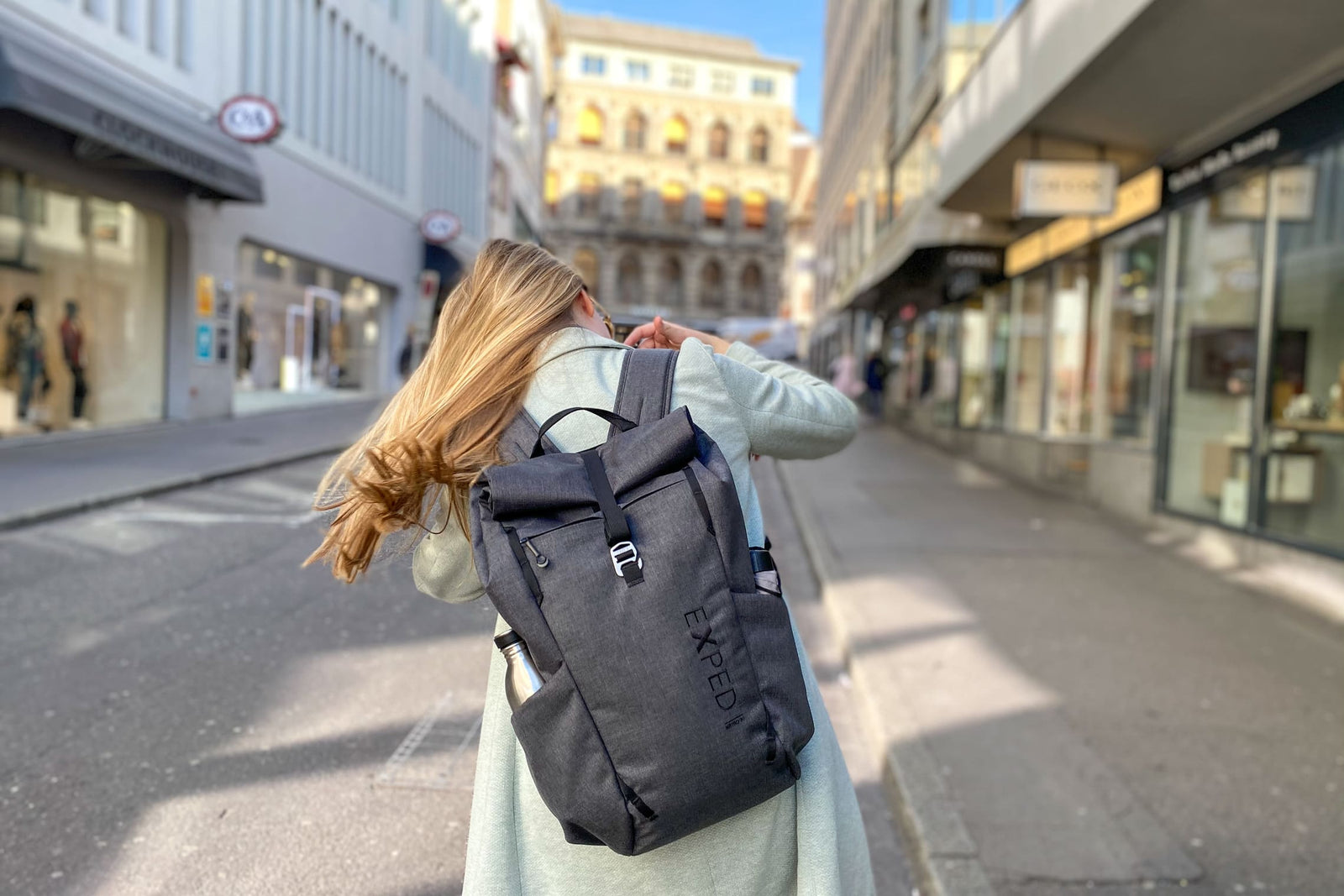 Eine Frau mit langen blonden Haaren, die einen hellgrauen Mantel trägt und einen EXPED Metro 30 Rucksack mit sich führt, geht eine sonnige, von Geschäften und Gebäuden gesäumte Stadtstraße entlang.