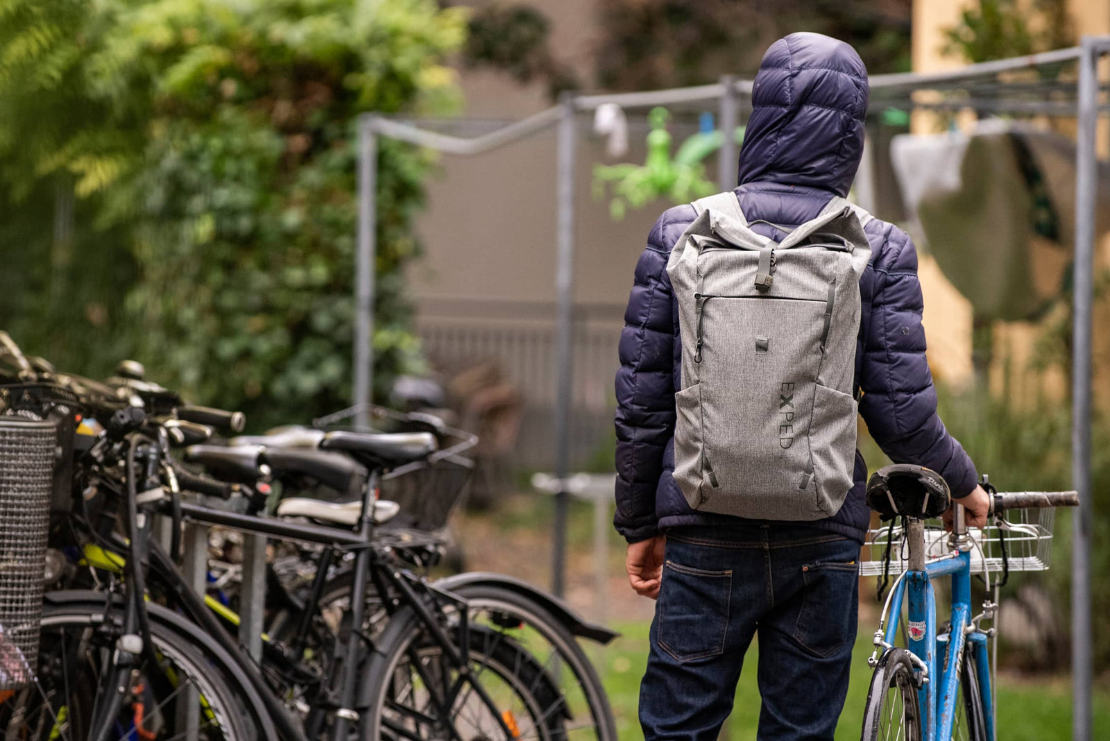 Eine Person in einer Kapuzenjacke steht an einem blauen Fahrrad, hält den Lenker fest und trägt einen EXPED Rucksack Metro 30 auf dem Rücken, in der Nähe von geparkten Fahrrädern im Freien im Grünen.