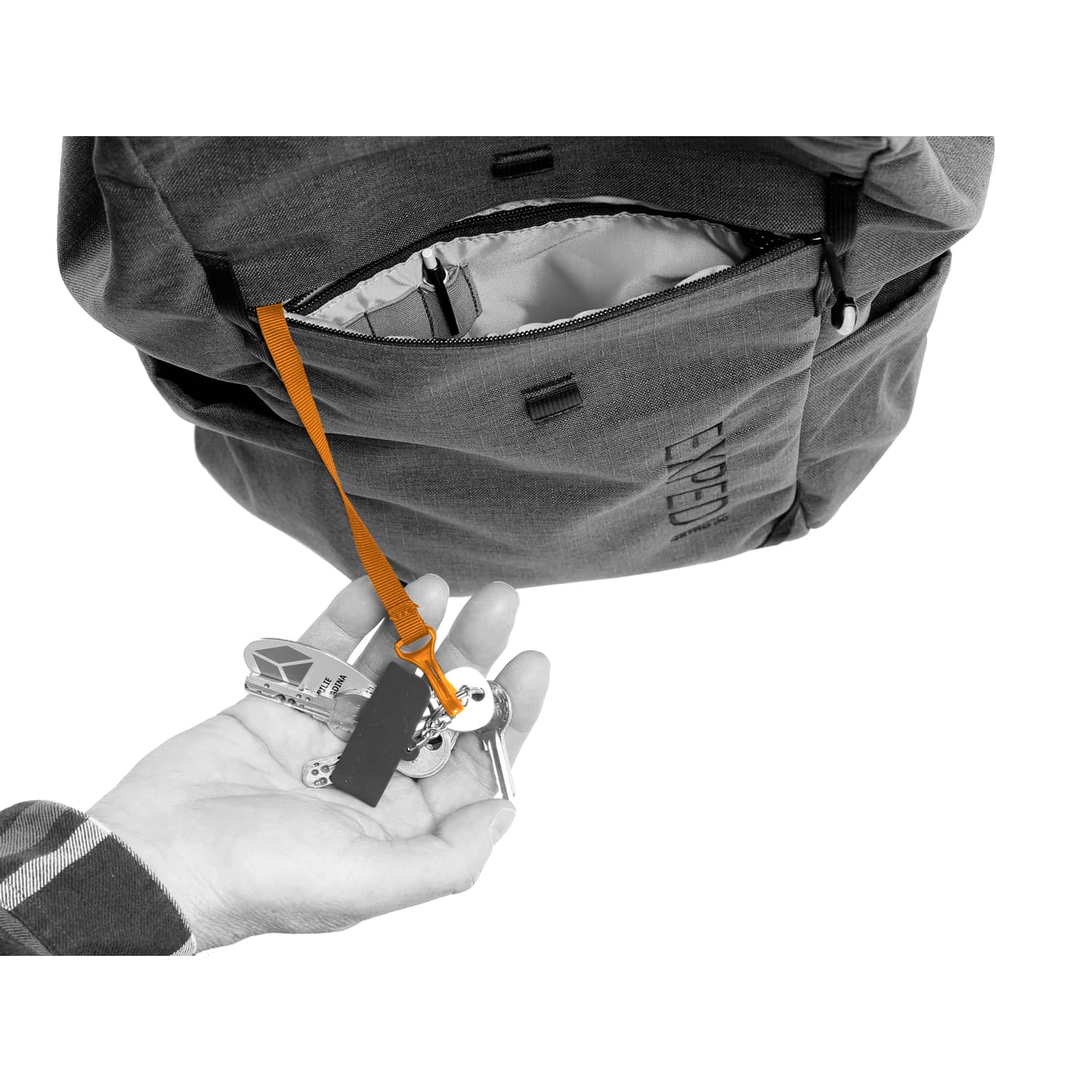 Eine Hand hält einen Schlüsselring, der an einem orangefarbenen Band befestigt ist, das in die Vordertasche des EXPED Rucksack Metro 30 von EXPED eingeklinkt ist. Die teilweise geöffnete Tasche gibt den Blick auf das helle Innere dieses urbanen Rucksacks frei.