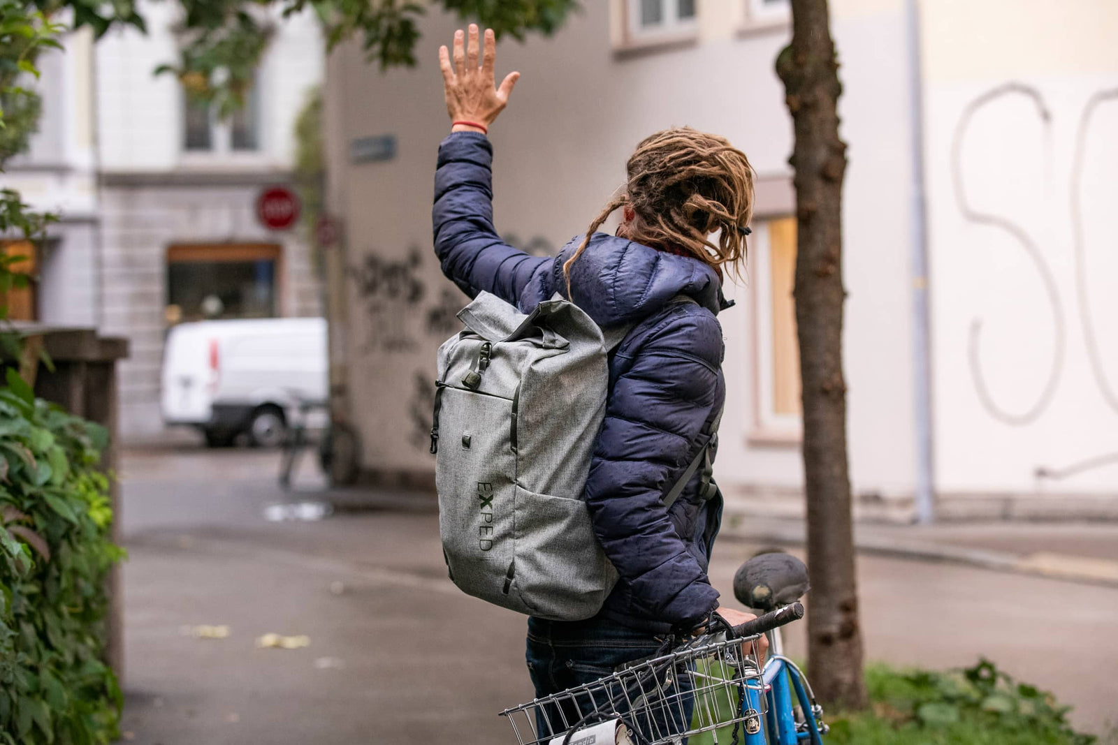 Eine Person mit Dreadlocks, bekleidet mit einer blauen Jacke und einem grauen EXPED Metro 30 Rucksack, steht neben einem Fahrrad und winkt auf einer von Gebäuden und Bäumen gesäumten Stadtstraße.