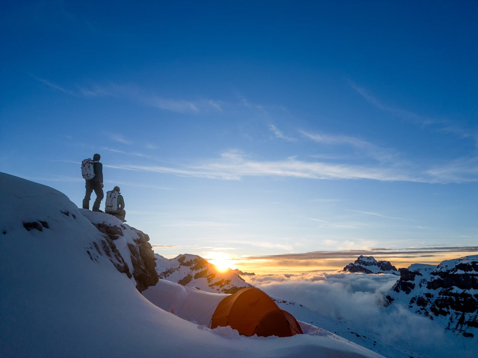 Zwei Personen mit Rucksäcken stehen bei Sonnenaufgang auf einem verschneiten Berg und blicken auf ihr EXPED Zelt Polaris. Weiche Wolken treiben am meist klaren Himmel, während ferne Gipfel in der aufgehenden Sonne glühen.