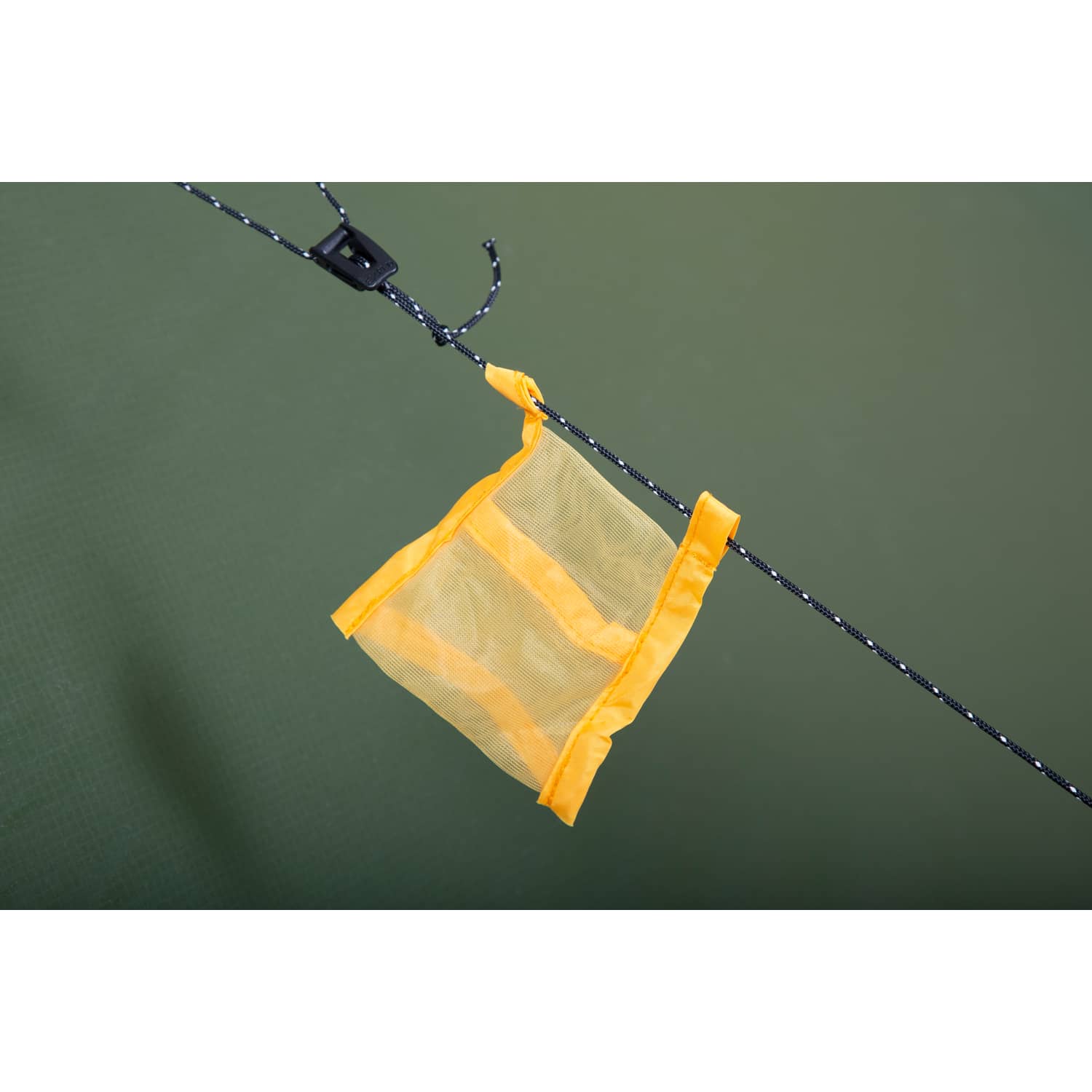 Eine kleine gelbe Netztasche, die an das Venus III DLX Extreme-Zubehör von EXPED erinnert, hängt an einer schwarz-weißen Kordel vor einem schlichten grünen Hintergrund.