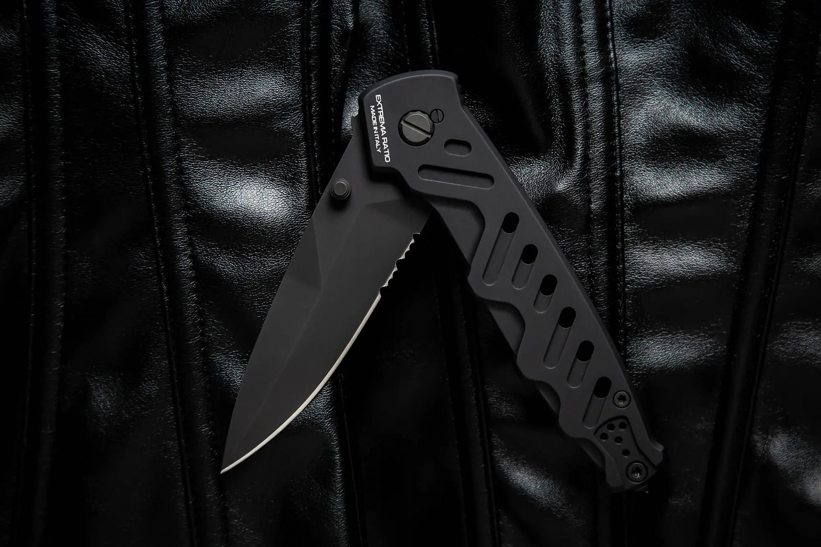 Ein EXTREMA RATIO Extrema Ratio Messer Caimano Nero N.A. mit teilgezahnter Klinge ist geöffnet und ruht auf einer glänzenden schwarzen Lederoberfläche mit sichtbaren Nähten.