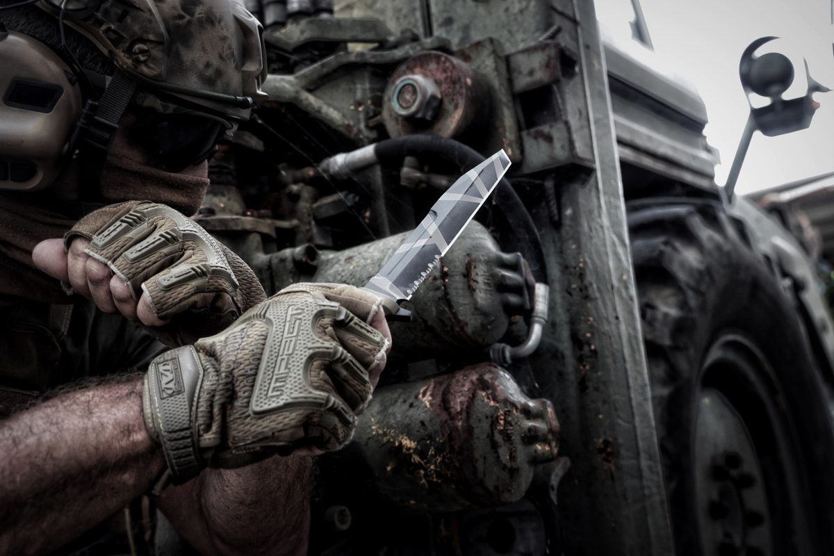 In taktischer Ausrüstung und mit Handschuhen greift eine Person ein EXTREMA RATIO Extrema Ratio Messer Fulcrum Desert Warfare Messer in der Nähe eines robusten Militärfahrzeugs und verkörpert so die Bereitschaft für ein hartes Überlebens- oder Kampfszenario.