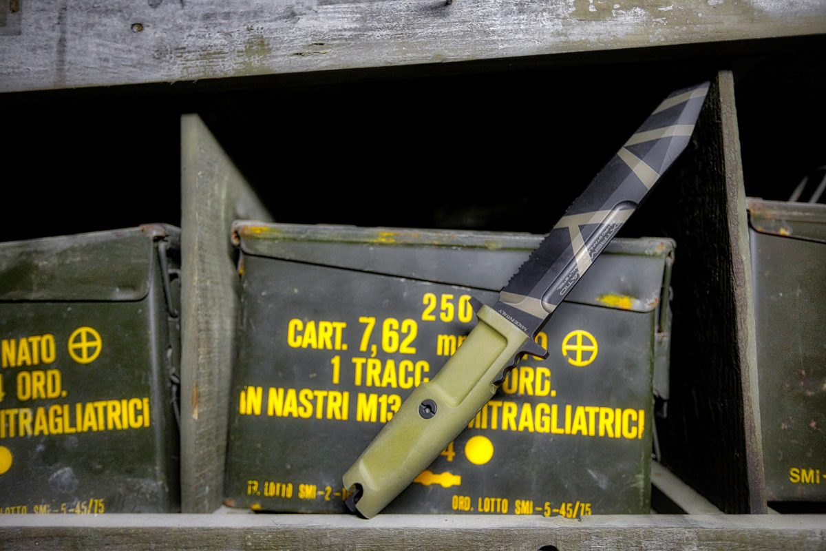 Ein EXTREMA RATIO Messer Fulcrum Desert Warfare stützt sich auf eine Munitionskiste aus Metall mit gelber Aufschrift, die auf einem verwitterten Holzregal ruht.