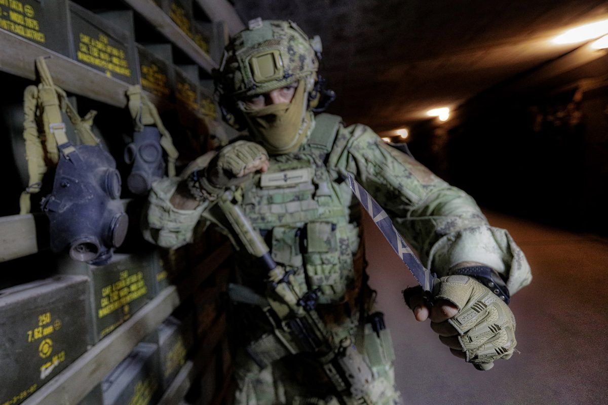 Ein Soldat in grünem Tarnanzug hält ein EXTREMA RATIO Messer Fulcrum Desert Warfare Messer defensiv in einem schummrigen Lagerraum, während im Hintergrund Kampfausrüstung und Gasmasken auf Regalen liegen.