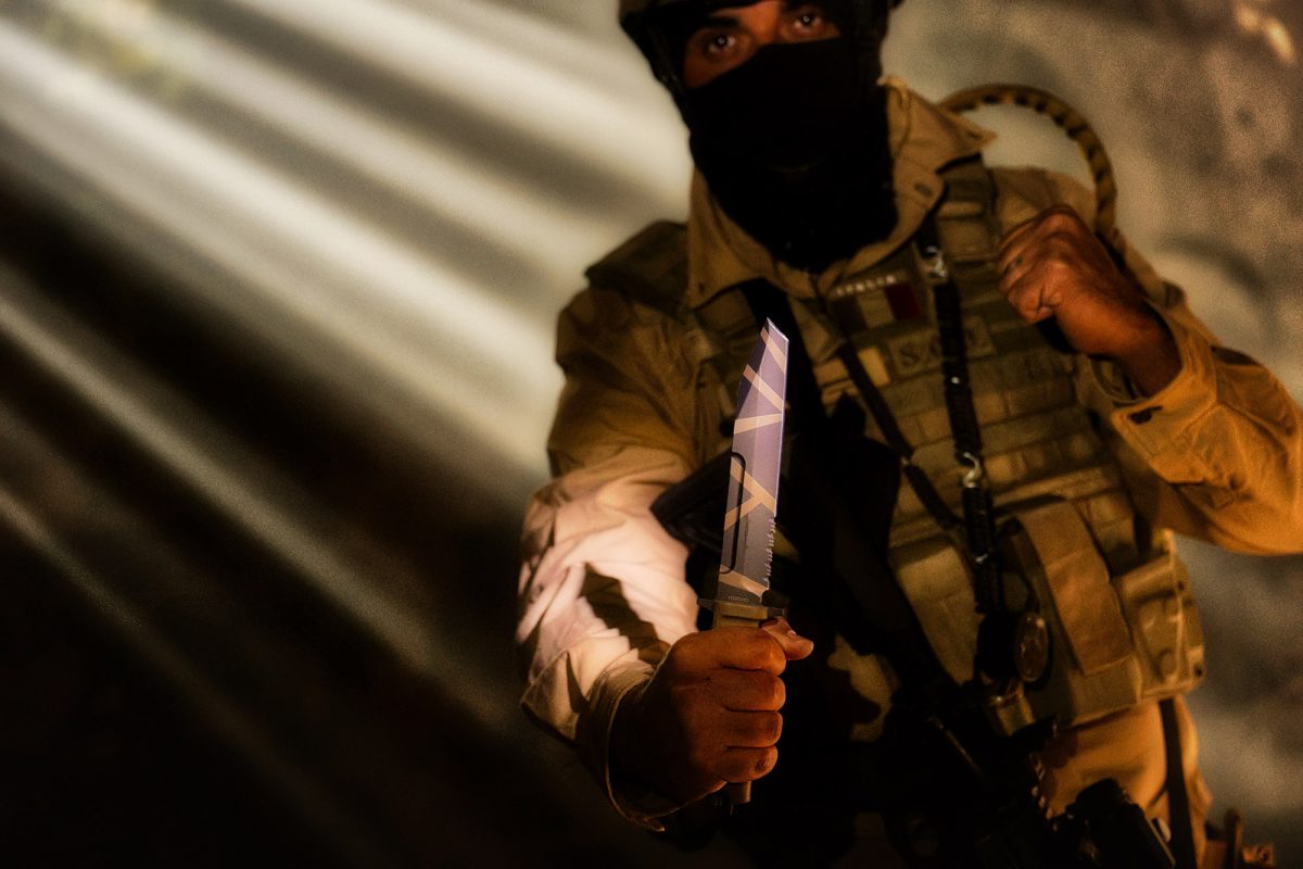 Eine Person in taktischer Ausrüstung und mit Gesichtsschutz streckt ein EXTREMA RATIO Messer Fulcrum Desert Warfare in Richtung Kamera, wobei dramatische Licht- und Schatteneffekte die intensive Stimmung noch verstärken.