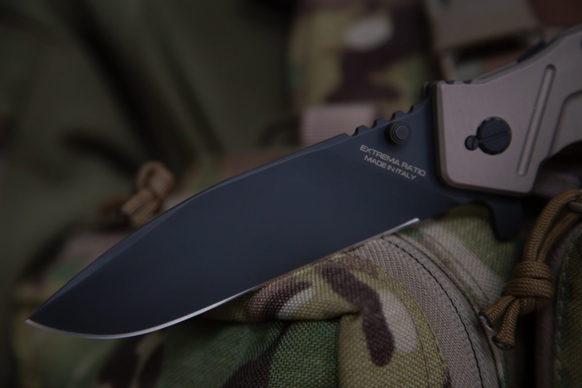 Eine Nahaufnahme des EXTREMA RATIO Extrema Ratio Messer Glauca G1 mit Böhler N690 Stahlklinge, abgebildet auf einem Stoff mit Tarnmuster und Ausrüstung in Weichzeichner.