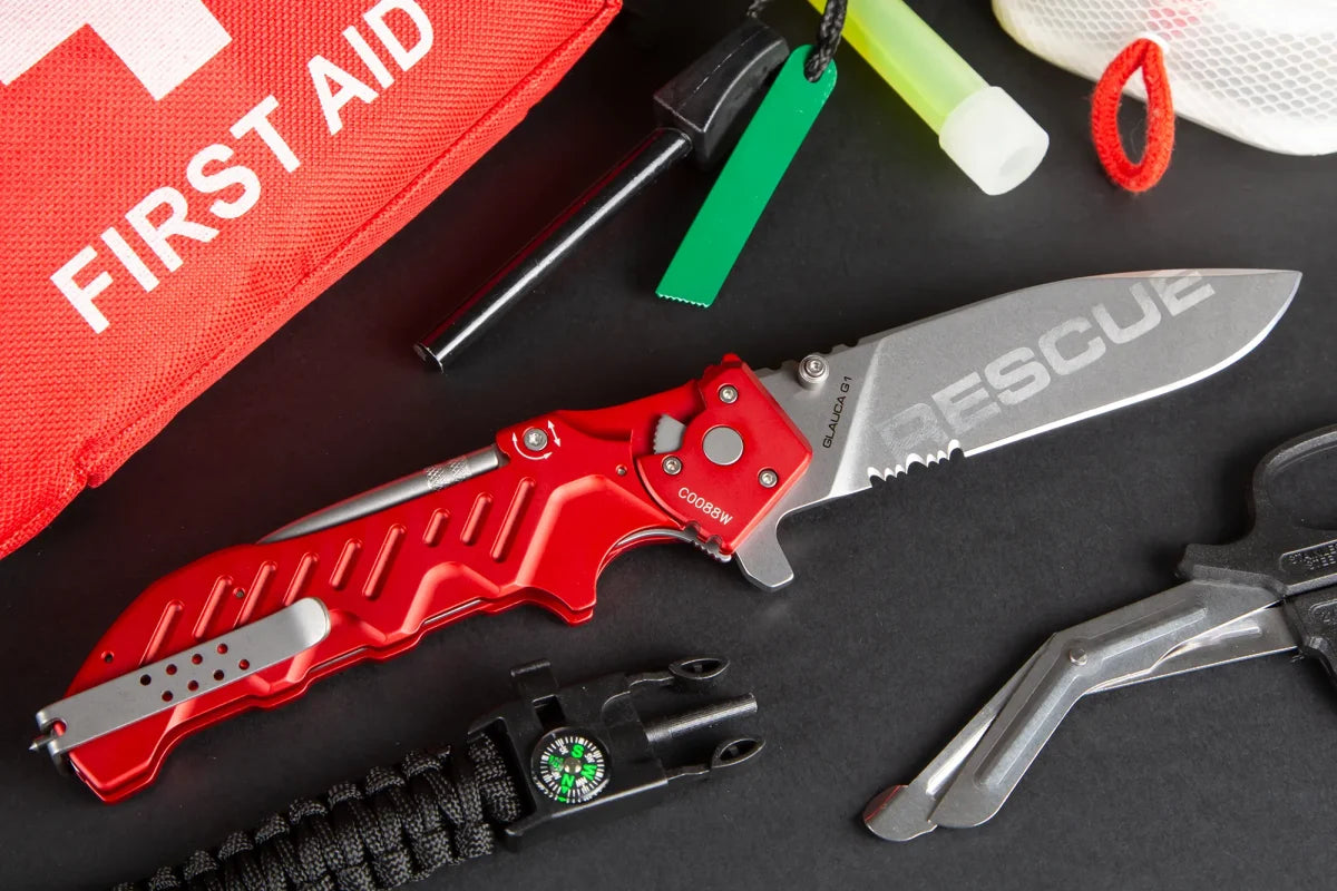 Ein EXTREMA RATIO Glauca G1 Rescue-Messer liegt neben einem roten Erste-Hilfe-Kasten, einem schwarzen Multi-Tool, einem Kompass-Armband, einer Pfeife und anderer Überlebensausrüstung auf einer schwarzen Fläche.