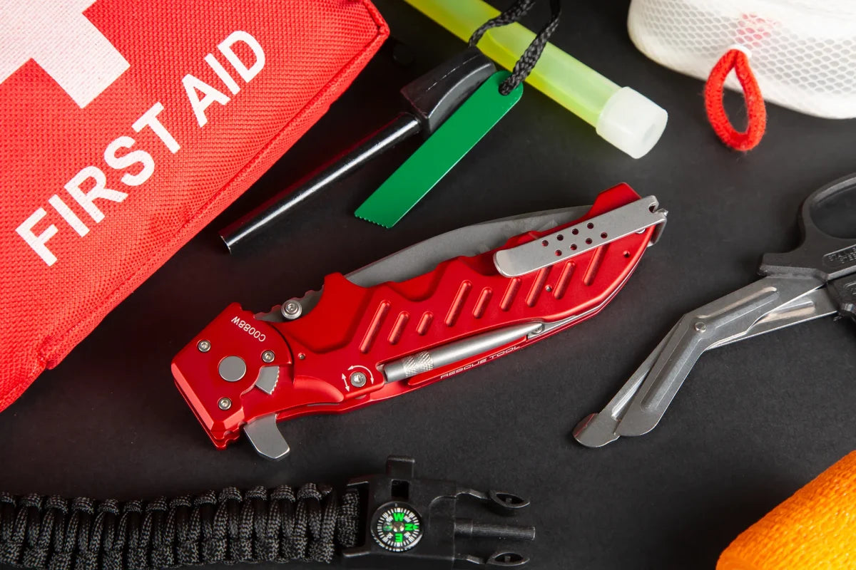 Auf einer schwarzen Fläche sind ein rotes Rettungsmesser, ein Erste-Hilfe-Set, eine Schere, eine Pfeife mit Kompass, ein Leuchtstab und andere Überlebensutensilien - darunter auch das EXTREMA RATIO Extrema Ratio Messer Glauca G1 Rescue - angeordnet.