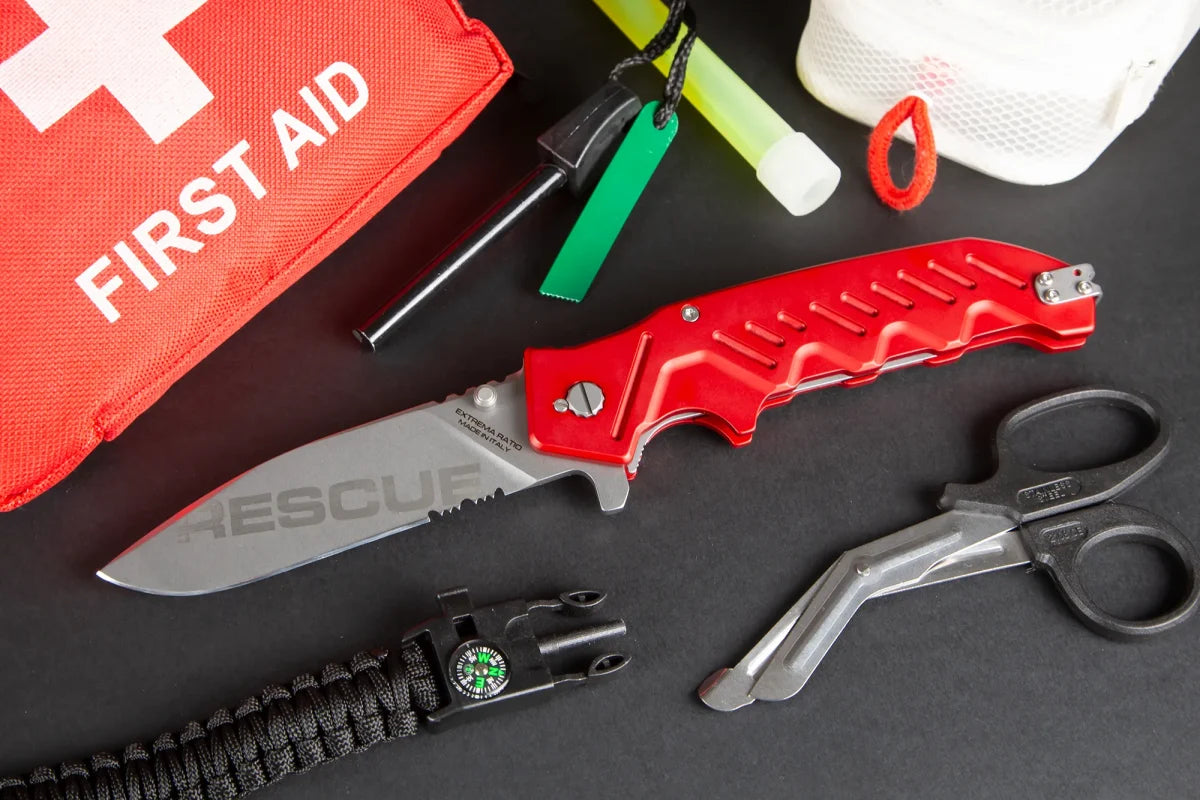 Ein EXTREMA RATIO Extrema Ratio Messer Glauca G1 Rescue Klappmesser (rot), eine Rettungsschere, ein Erste-Hilfe-Set, ein Paracord-Armband mit Kompass, ein Leuchtstab und eine weiße Netztasche sind auf einer schwarzen Fläche ausgestellt.