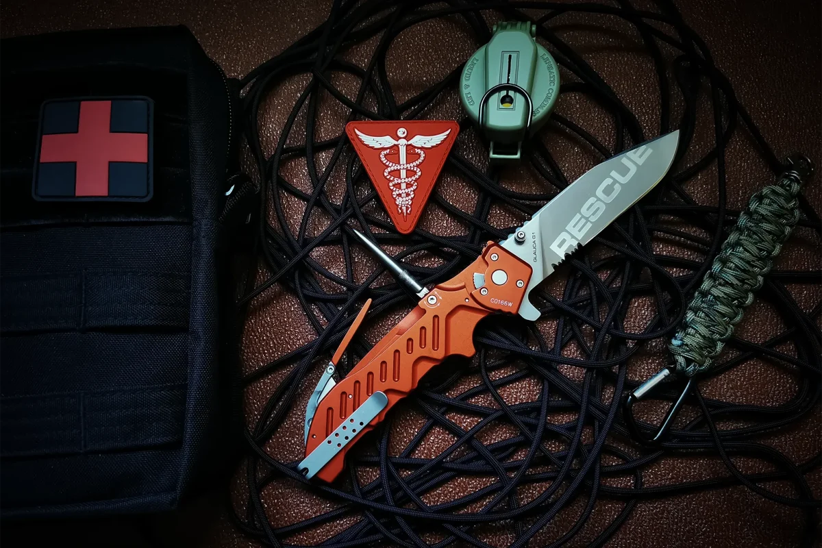 Ein Rettungsmesser, ein medizinisches Pflaster, ein Caduceus-Emblem, ein Kompass, ein Paracord und ein aufgerolltes Seil liegen wie ein Notfallset auf einer dunklen Fläche. Das EXTREMA RATIO Extrema Ratio Messer Glauca G1 Rescue zeichnet sich durch taktische Effizienz aus.
