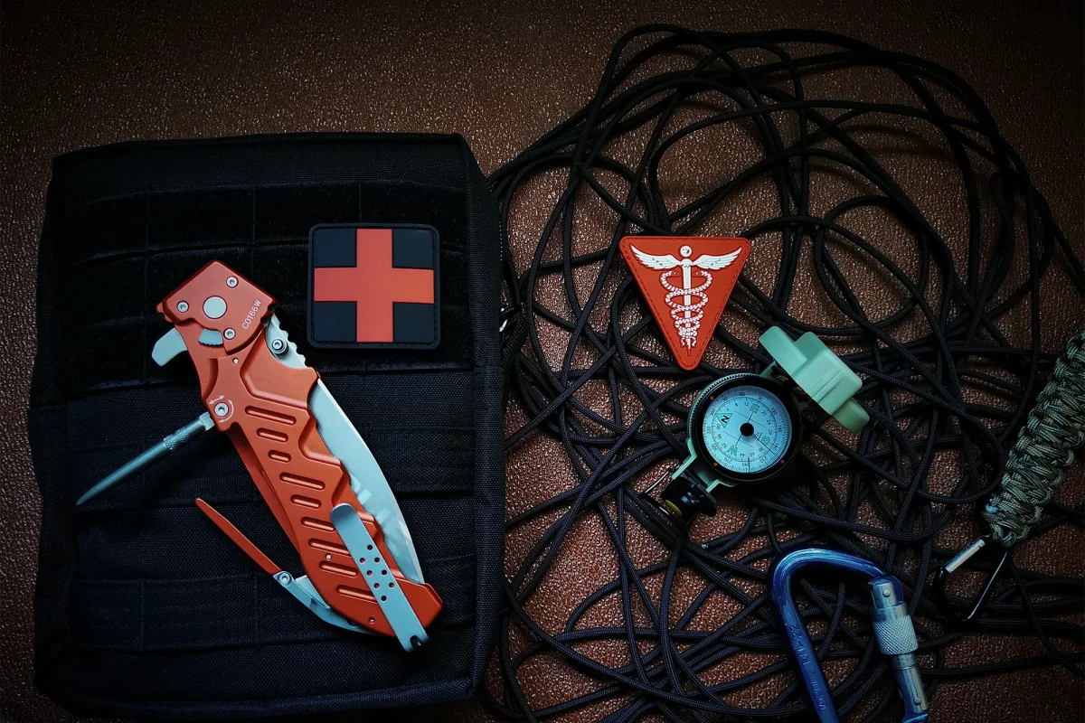 Eine taktische Tasche mit rotem Sanitätskreuz, orangefarbenem Extrema Ratio Messer Glauca G1 Rescue von EXTREMA RATIO, Caduceus-Patch, Stethoskop, schwarzer Kordel und Paracord-Armband sind auf einer braunen Fläche angeordnet.