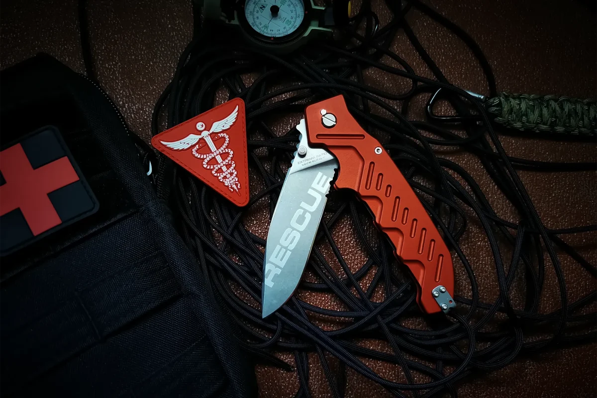 Ein Extrema Ratio Messer Glauca G1 Rescue Klappmesser von EXTREMA RATIO, mit "RESCUE" auf der Klinge, ruht auf schwarzen Kordeln neben einem Erste-Hilfe-Kasten und einem roten dreieckigen Aufnäher mit dem medizinischen Symbol des Caduceus.