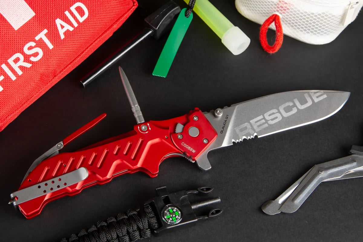 Ein EXTREMA RATIO Extrema Ratio Messer Glauca G1 Rescue mit teilweise gezahnter Klinge liegt zwischen Notfallwerkzeugen, einem Erste-Hilfe-Set, einer Pfeife und einem Paracord-Armband mit Kompass auf einer schwarzen Oberfläche.