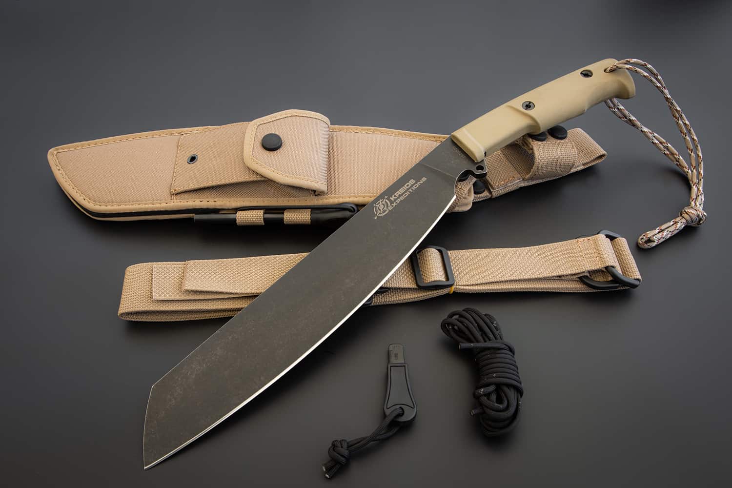 Das EXTREMA RATIO Messer Kreios Expedition, ein großes Überlebensmesser mit hellbraunem Griff und schwarzer Klinge, wird in einer passenden hellbraunen Scheide mit Riemen gezeigt, zusammen mit einer schwarzen Kordel und einem kleinen Werkzeug, alles auf einer dunklen Oberfläche.