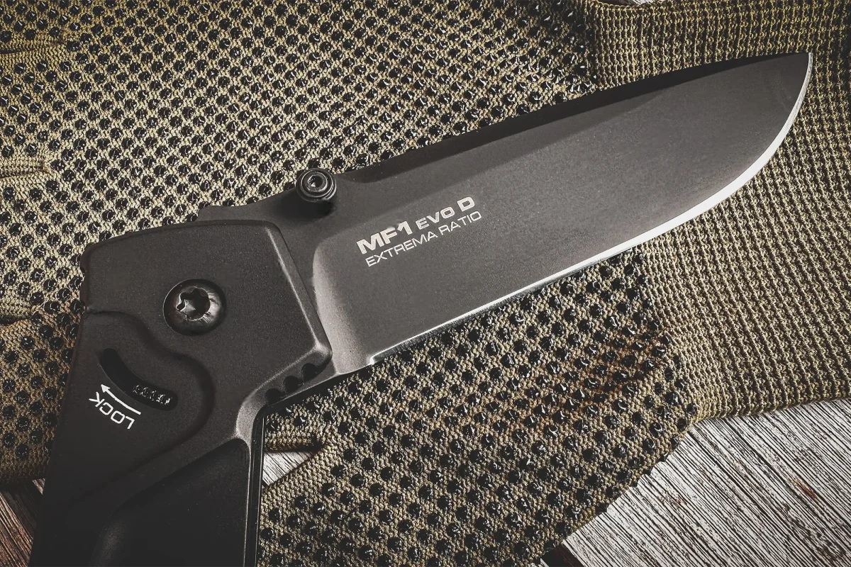 Das EXTREMA RATIO Messer MF1 EVO D Black verfügt über eine schwarze Tanto-Klinge von Böhler M390 und einen taktischen Griff, der offen auf einem olivgrünen, strukturierten Stoff abgebildet ist.