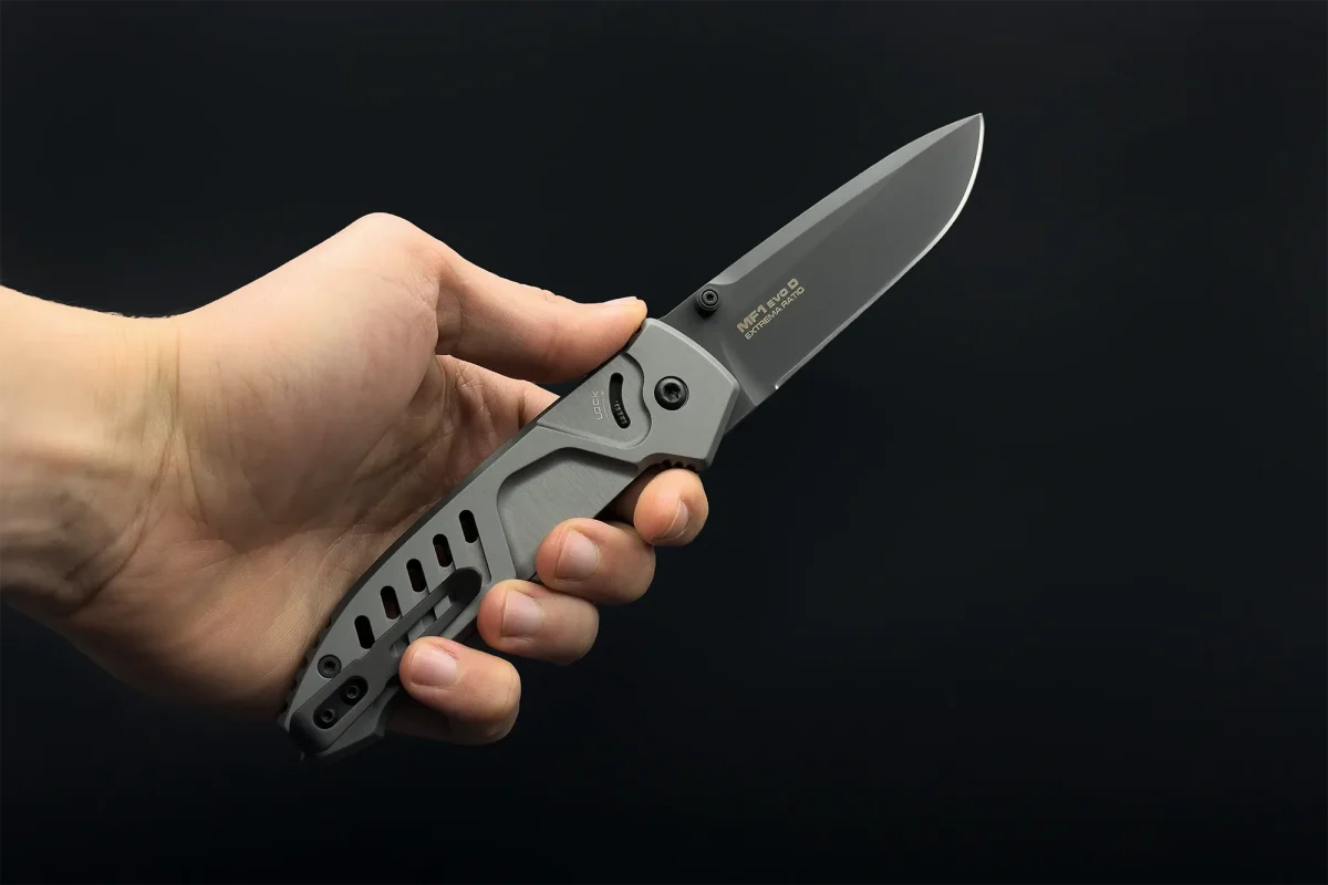 Eine Hand hält ein geöffnetes EXTREMA RATIO Extrema Ratio Messer MF1 EVO D Tactical Grey Klappmesser mit Metallgriff und schwarzer Böhler N690 Klinge vor einem schlichten schwarzen Hintergrund.