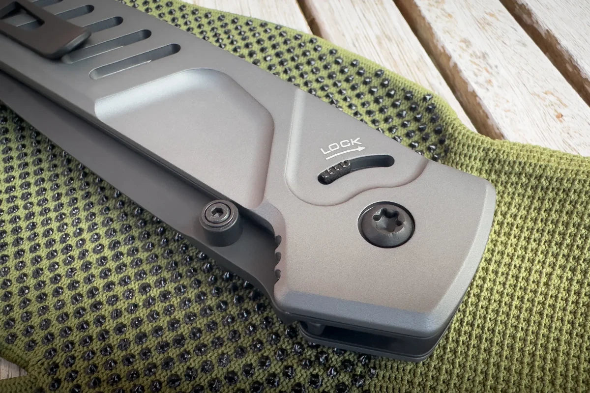 Eine Nahaufnahme des EXTREMA RATIO Extrema Ratio Messer MF1 EVO D Tactical Grey mit Böhler N690 Stahl hebt den Verschlussmechanismus hervor, während es auf einem Holztisch auf grünem Strukturstoff liegt.