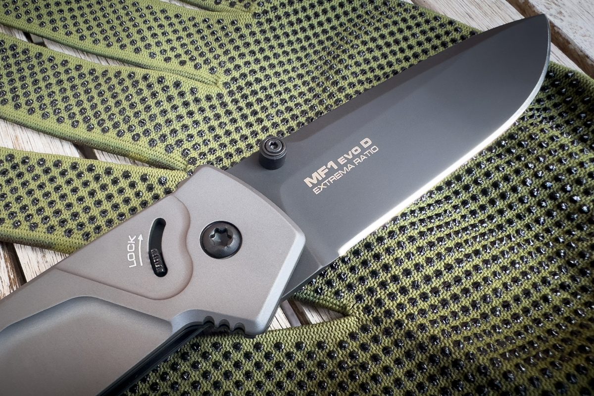 Eine Nahaufnahme des EXTREMA RATIO Extrema Ratio Messer MF1 EVO D Tactical Grey Klappmessers aus Böhler N690 Stahl auf grün-schwarzen Handschuhen auf einer Holzoberfläche.