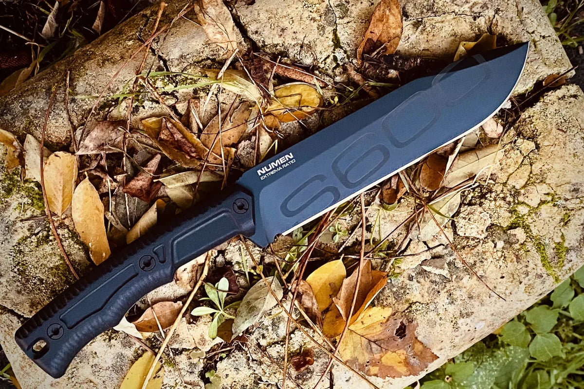 Ein Extrema Ratio Messer Numen S600 Black von EXTREMA RATIO ruht auf einem Felsen zwischen trockenen Blättern und Zweigen. Sein strukturierter Griff und die Klinge aus Böhler S600-Stahl, die mit geometrischen Mustern "NUVEM" gekennzeichnet ist, heben sich von der Szene ab.