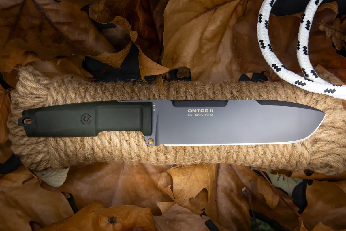Das Extrema Ratio Messer Ontos II S600 Black, ein feststehendes Messer mit schwarzem Griff und EXTREMA RATIO Branding auf der Klinge, ist auf einer Seilspule zwischen braunen Herbstblättern und einer weiß-schwarz gemusterten Kordel platziert.