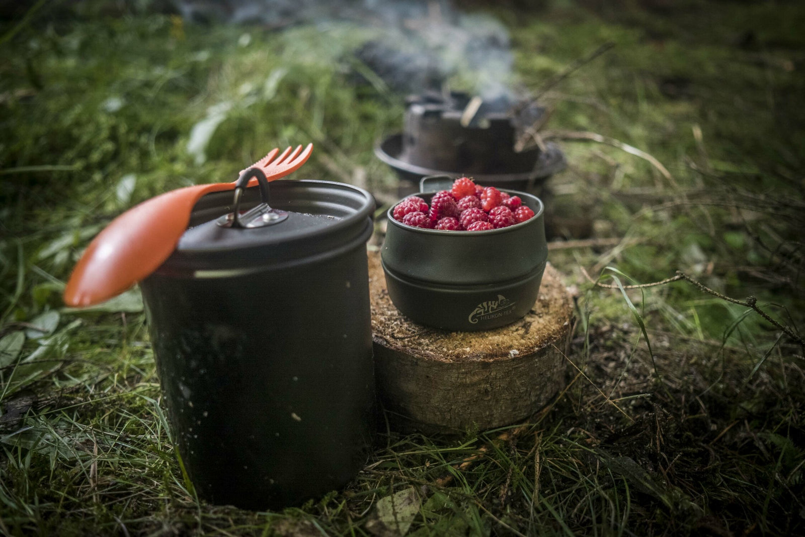 Auf der Wiese ist dunkles Camping-Kochgeschirr zu sehen: ein Topf mit einem orangefarbenen Göffel auf dem Deckel, der HELIKON-TEX Helikon-Tex Camp Cup, gefüllt mit frischen Himbeeren auf einer Holzscheibe, und ein rauchender Campingkocher im Hintergrund.