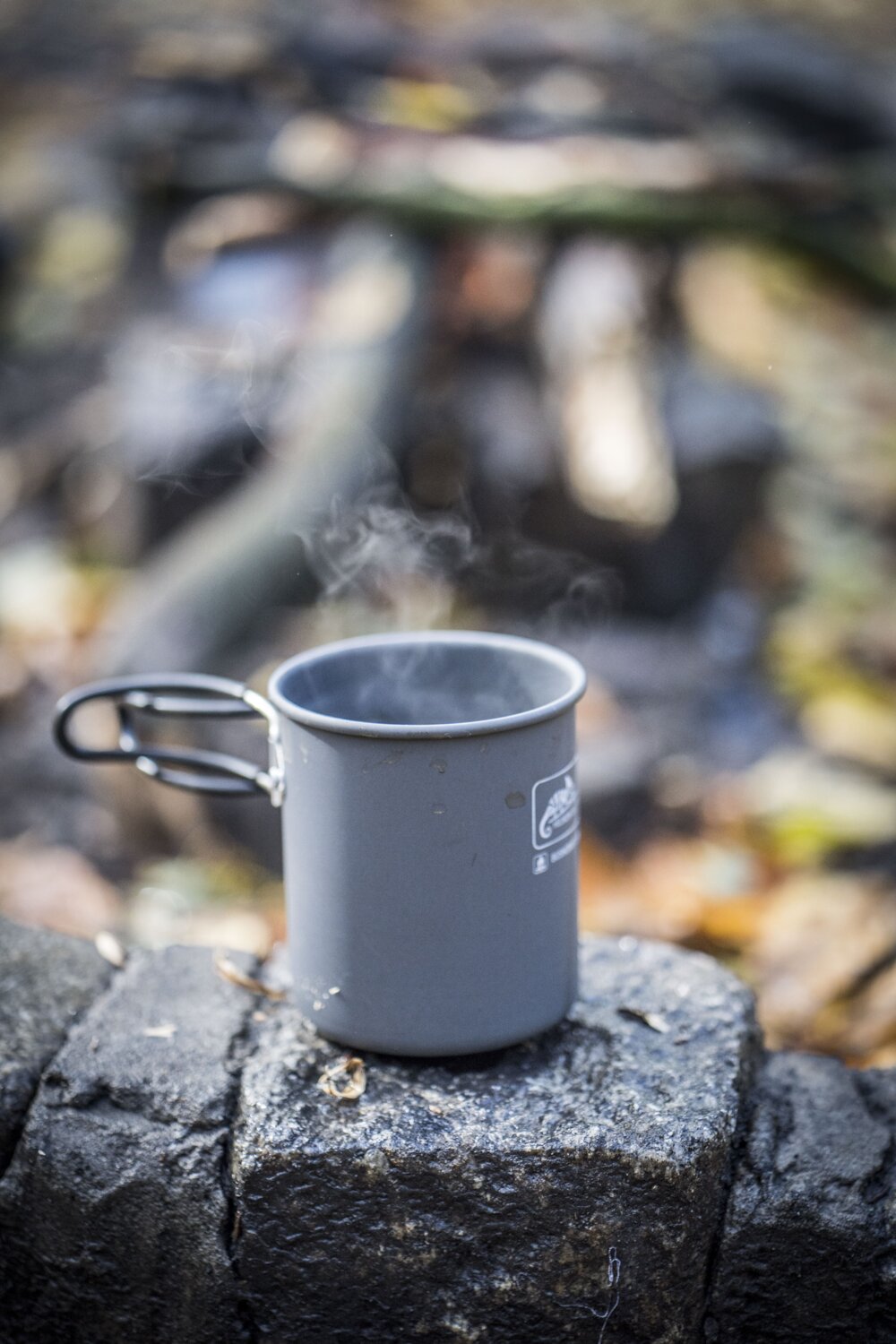 Ein HELIKON-TEX Camp Cup steht auf einem Stein im Freien, mit einem verschwommenen Hintergrund aus Blättern und Ästen, der an den Genuss eines warmen Getränks in der Natur erinnert.