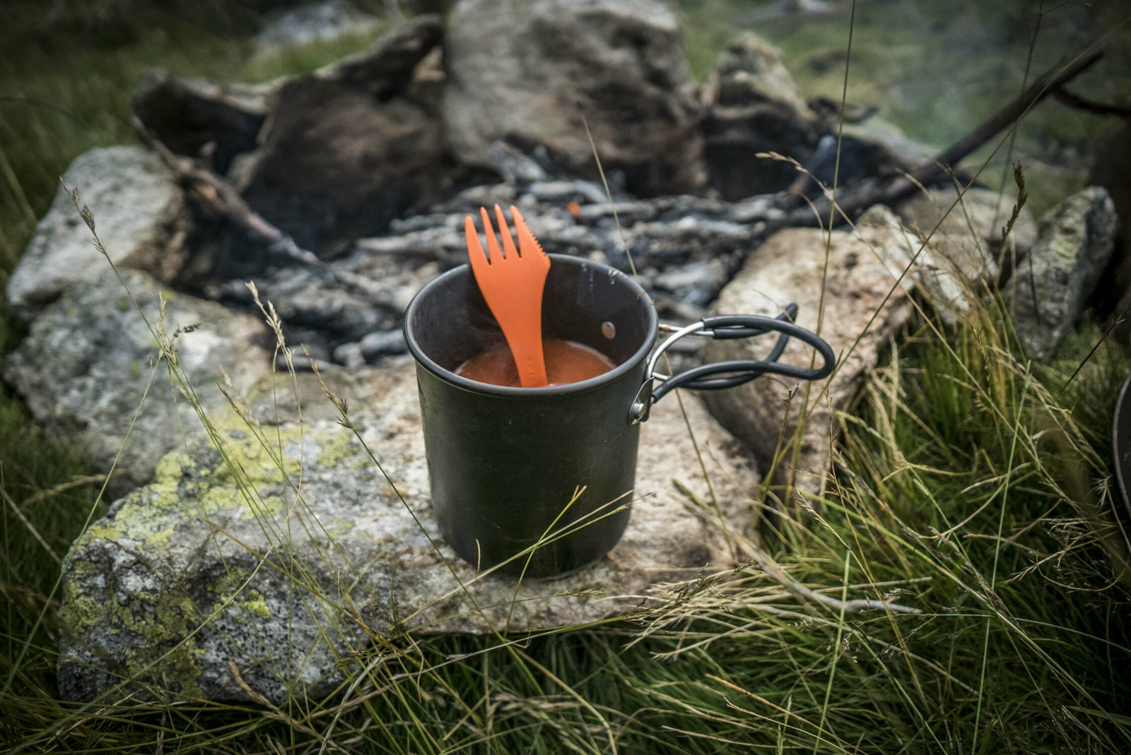 Ein dunkler HELIKON-TEX Helikon-Tex Camp Cup und eine orangefarbene Plastikgabel ruhen auf einem Felsen in der Nähe eines steinernen Lagerfeuers, umgeben von Gras und Steinen im Freien.