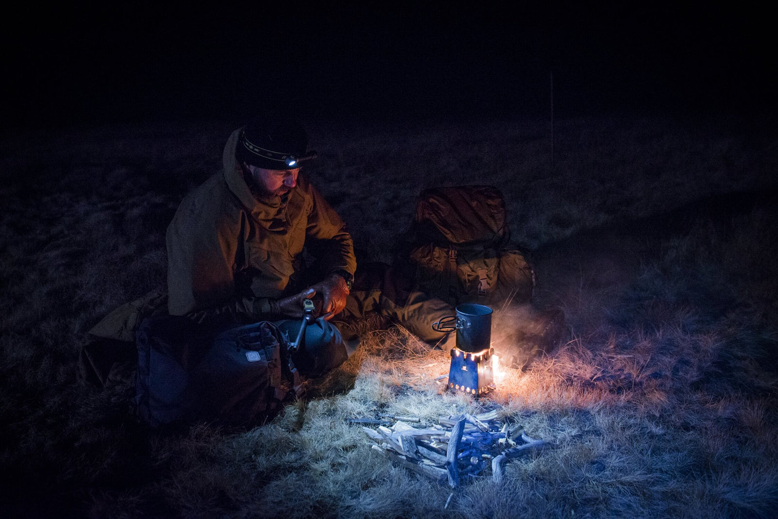 Ein Camper in Outdoor-Ausrüstung und mit Stirnlampe kümmert sich nachts um ein kleines Lagerfeuer und einen Kocher. Der HELIKON-TEX Helikon-Tex Camp Cup steht neben dem Rucksack und wird vom Feuerschein auf dem trockenen Gras beleuchtet.