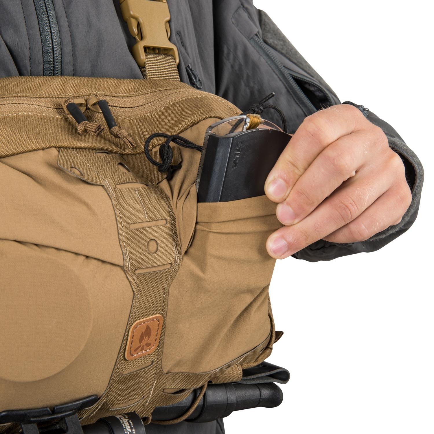 Eine Person in einer grauen Jacke packt einen schwarzen Gegenstand in den HELIKON-TEX Chest Pack Numbat® - Earth Brown / Clay B, indem sie dessen praktische Fronttasche während eines Outdoor-Abenteuers benutzt.