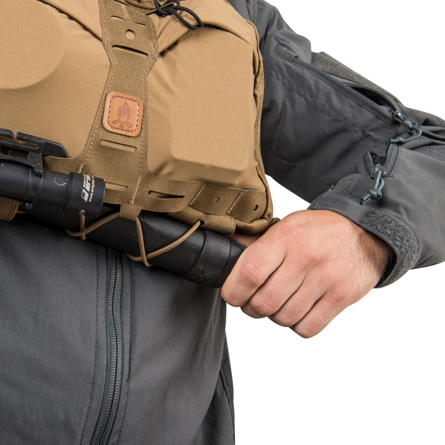 Eine Person in einer grauen taktischen Jacke zieht eine schwarze Taschenlampe oder einen Schlagstock aus dem unteren MOLLE-Gurt eines HELIKON-TEX Chest Pack Numbat® in Earth Brown / Clay B.
