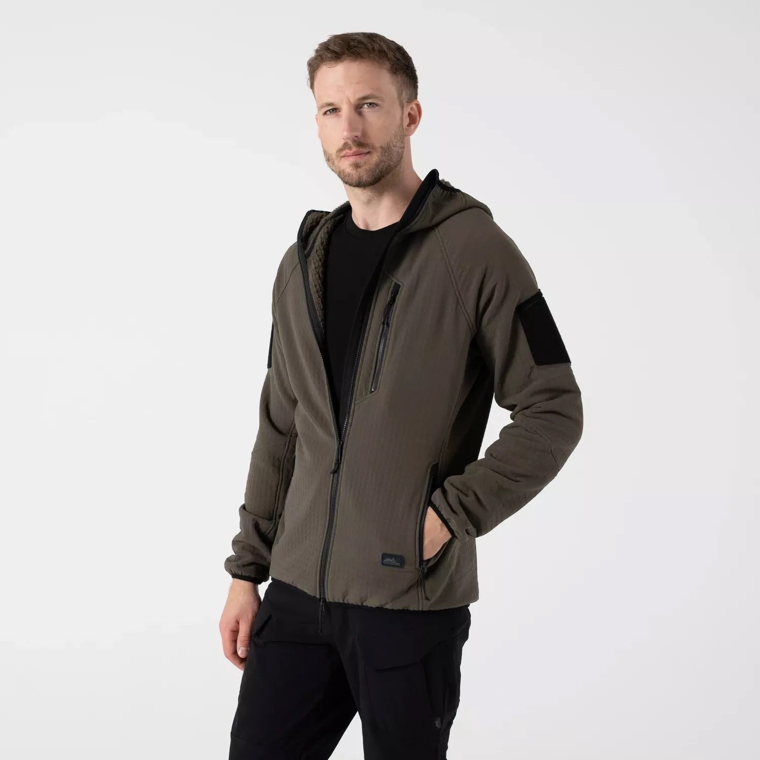 Ein Mann mit kurzen Haaren und Bart trägt das HELIKON-TEX Patriot PRO Fleece in Taiga Green mit schwarzen Akzenten und passender schwarzer Hose, stehend vor einem schlichten weißen Hintergrund.