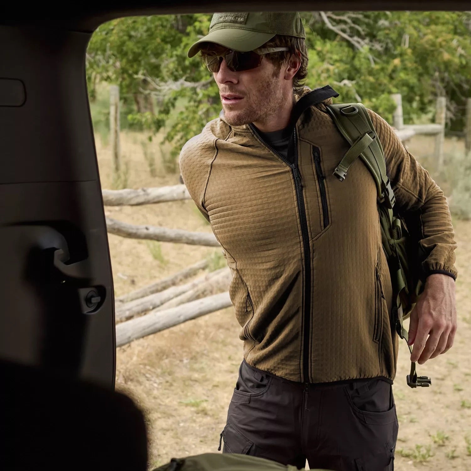 Ein Mann mit Sonnenbrille und Mütze, bekleidet mit einem HELIKON-TEX Patriot PRO Fleece - Taiga Green, steht an einer offenen Autotür in der Nähe von Bäumen und einem Holzzaun und setzt einen grünen Rucksack auf.