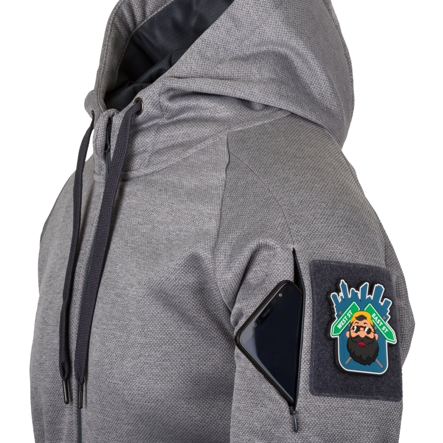 Der Helikon-Tex Urban Tactical Hoodie (FullZip)® - Melange Black-Grey von HELIKON-TEX hat dunkle Kordelzüge, einen grauen Körper, eine Ärmeltasche mit Reißverschluss, in der ein schwarzes Smartphone steckt, und einen bunten Aufnäher mit einem bärtigen Mann mit Sonnenbrille und Hut.