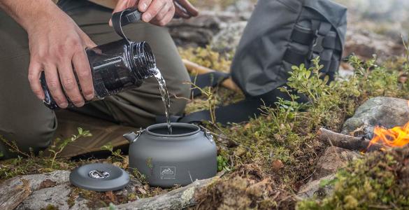 Ein Camper gießt Wasser aus einer schwarzen Flasche in den HELIKON-TEX Wasserkessel Camp Kettle neben einem kleinen Lagerfeuer, mit Gras, Moos, Steinen und einem grauen Rucksack in der Nähe - ideal für Bushcraft-Abenteuer.
