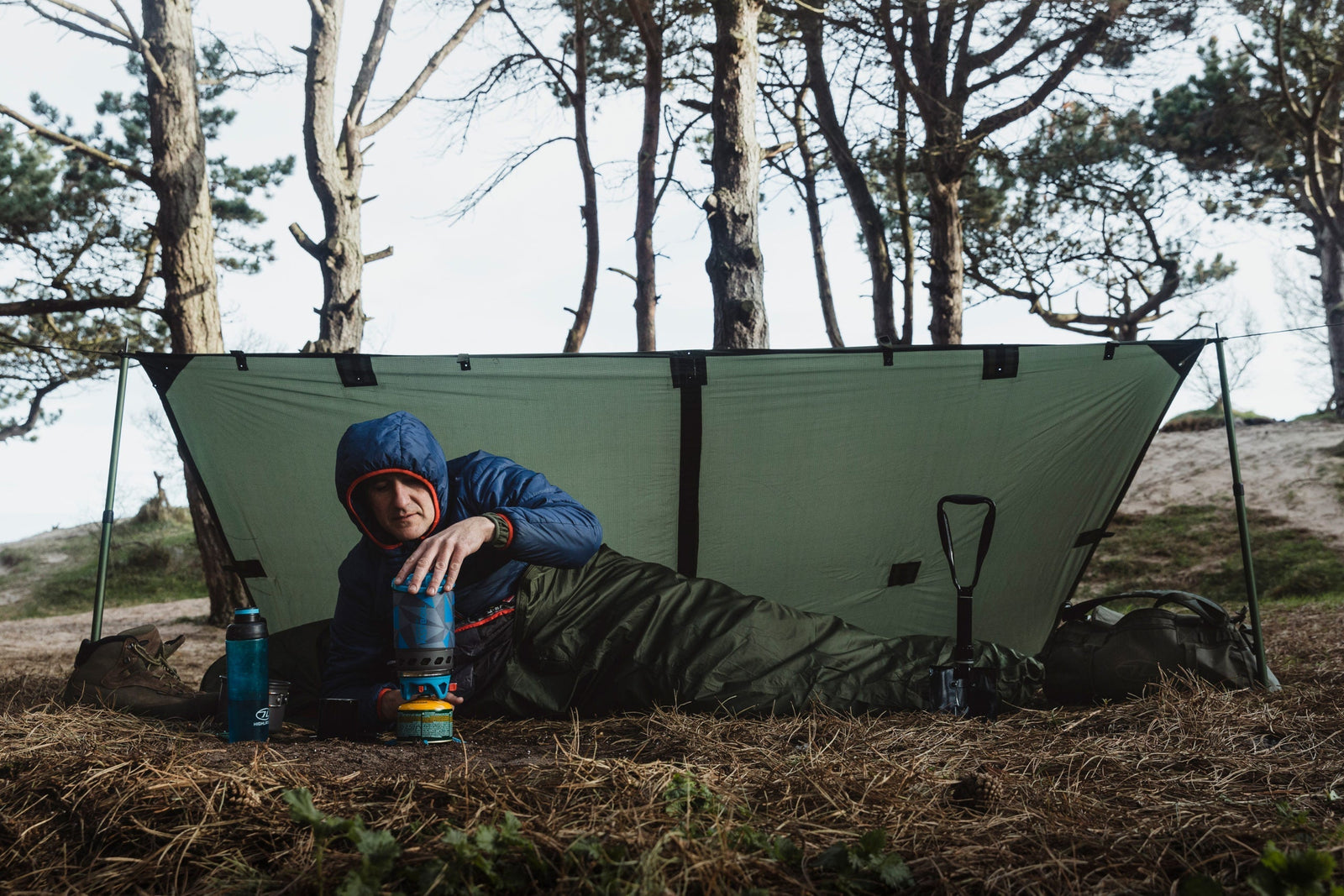 Eine Person in Outdoor-Kleidung liegt in einem HIGHLANDER FORCES Highlander Biwaksack Hawk Bivy Bag unter einer grünen Plane in einem Wald und benutzt einen Campingkocher. In der Nähe befinden sich eine Schaufel und Wasserflaschen, mit Bäumen und sandigem Boden im Hintergrund.