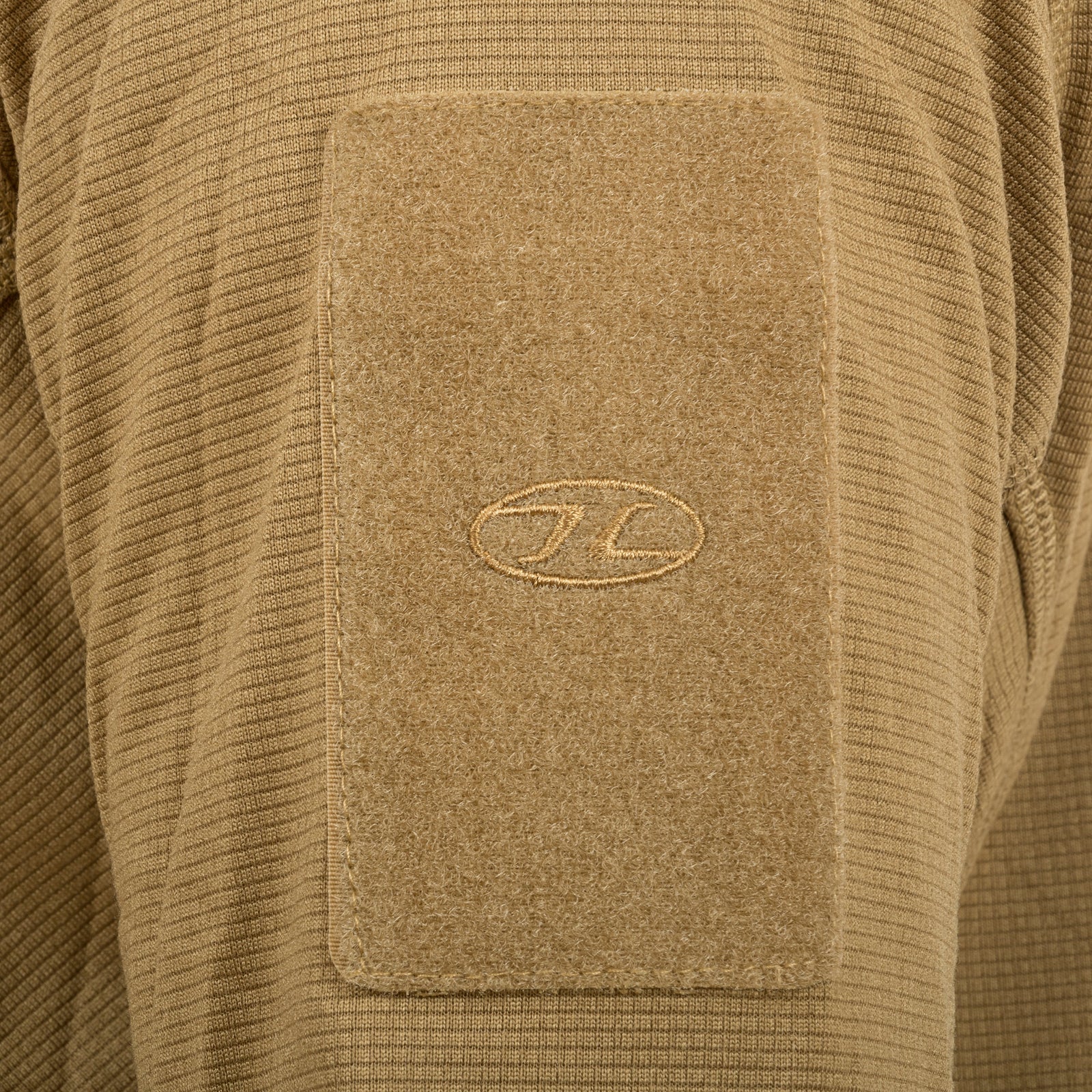 Nahaufnahme eines Stoffaufnähers in Coyote Tan mit einem dezenten ovalen Logo, der auf dem Ärmel eines HIGHLANDER FORCES Tactical Hirta Fleece - Coyote Tan - auf einem passenden gerippten Textilhintergrund angebracht ist.