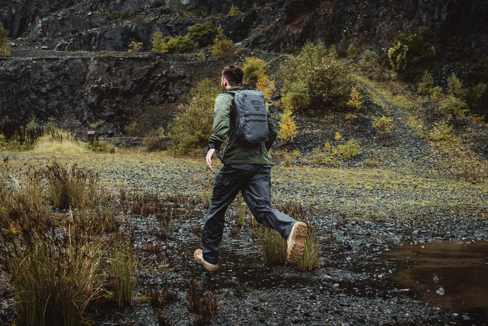 Mit einem HIGHLANDER FORCES Tactical Hirta Fleece - Coyote Tan wandert ein Mann mit Rucksack an einem bewölkten Tag durch felsiges, nasses Gelände, das mit Gras und entfernten Bäumen übersät ist.