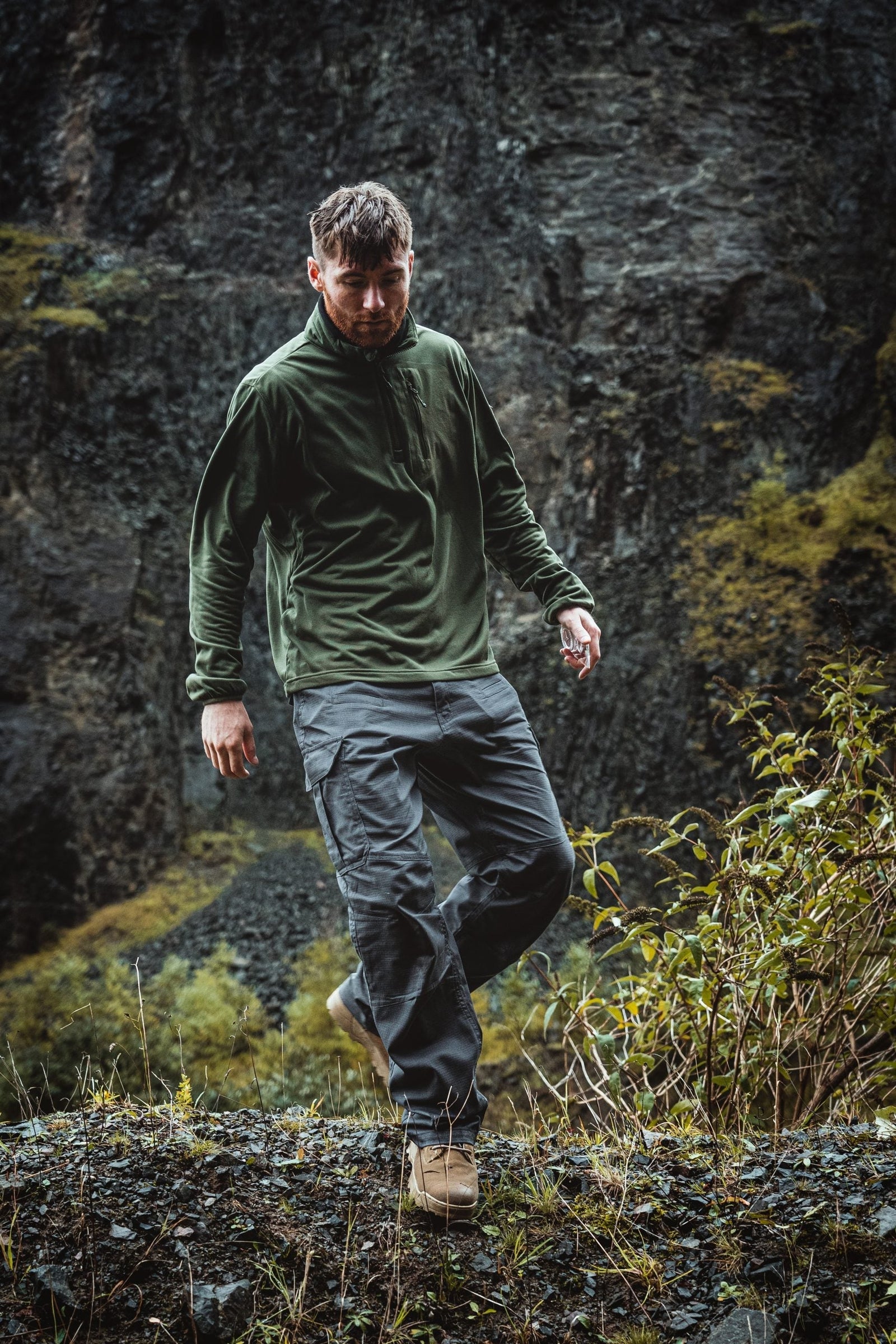 Ein Mann in einem HIGHLANDER FORCES Tactical Hirta Fleece - Ranger Green und einer dunklen Hose geht im Freien auf felsigem Gelände, mit einem steilen Abhang hinter ihm und grüner Vegetation im Vordergrund.