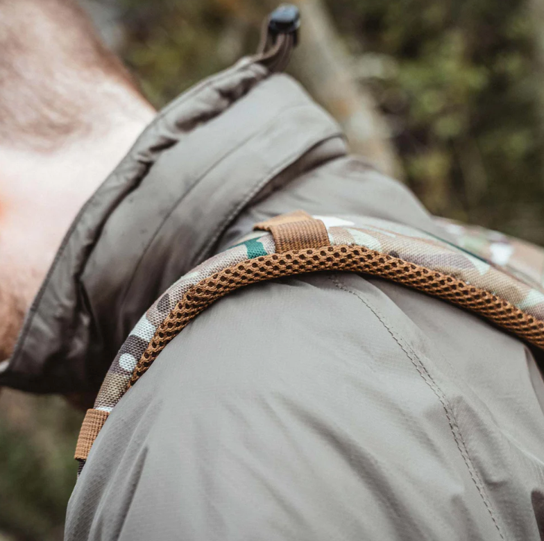 Nahaufnahme der Schulter einer Person, die einen schwarzen Highlander Halo Full Zip Tactical Smock von HIGHLANDER FORCES mit einem tarnfarbenen, winddichten und wasserabweisenden Rucksackträger und hellbrauner Netzpolsterung trägt, im Freien mit unscharfem grünen Laub.